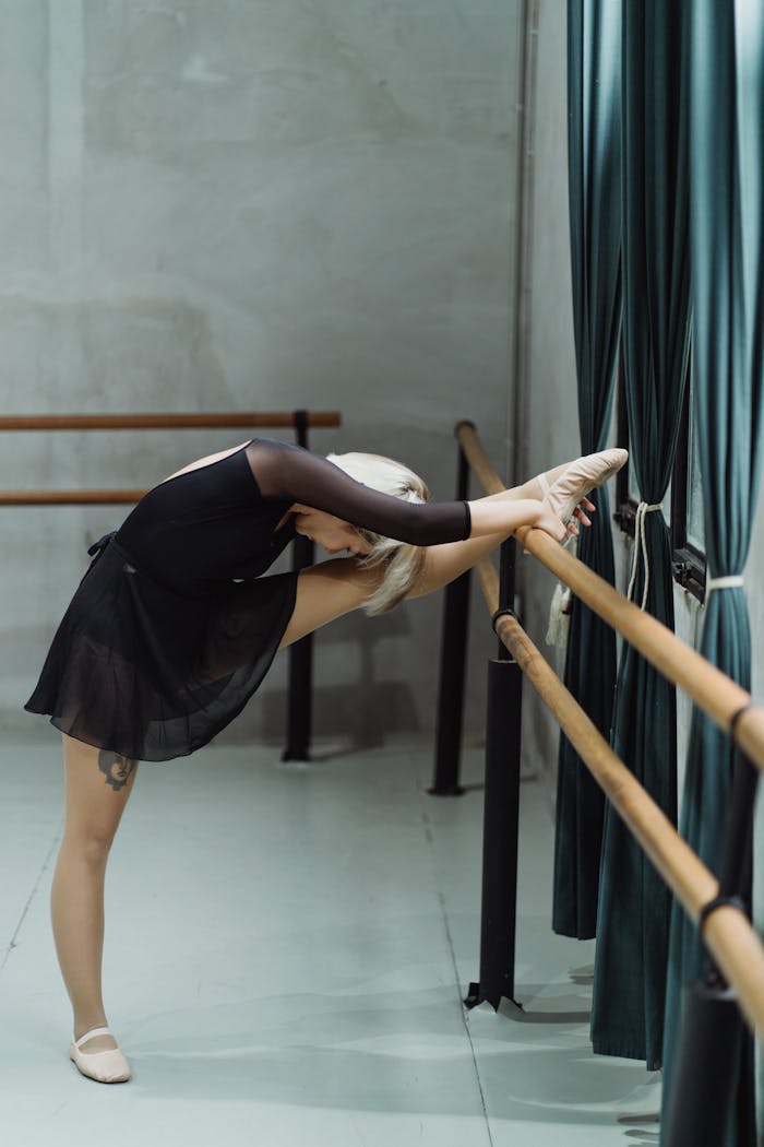 A graceful ballerina in a dark dress stretches at the barre in a dance studio, showcasing flexibility and elegance.